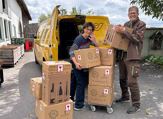 Eva und Norbert Eder vom Pankrazhofer stehen neben einem gelben Post-Transporter und stapeln Pakete mit Glas-Warnaufklebern auf einem Rollwagen. Sie lächeln und winken in die Kamera.