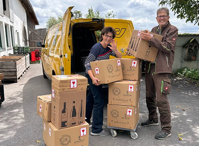 Eva und Norbert Eder vom Pankrazhofer stehen neben einem gelben Post-Transporter und stapeln Pakete mit Glas-Warnaufklebern auf einem Rollwagen. Sie lächeln und winken in die Kamera.