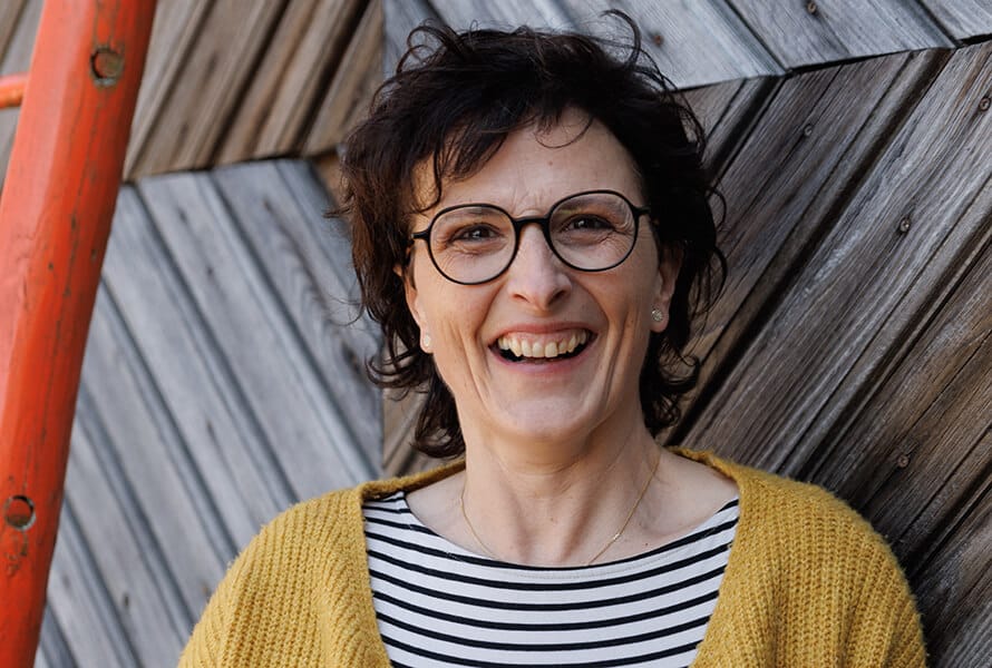 Eva Eder vom Pankrazhofer lächelt in die Kamera. Sie trägt Brille, einen gestreiften Pullover und einen senfgelben Cardigan. Im Hintergrund eine diagonale Holzfassade.
