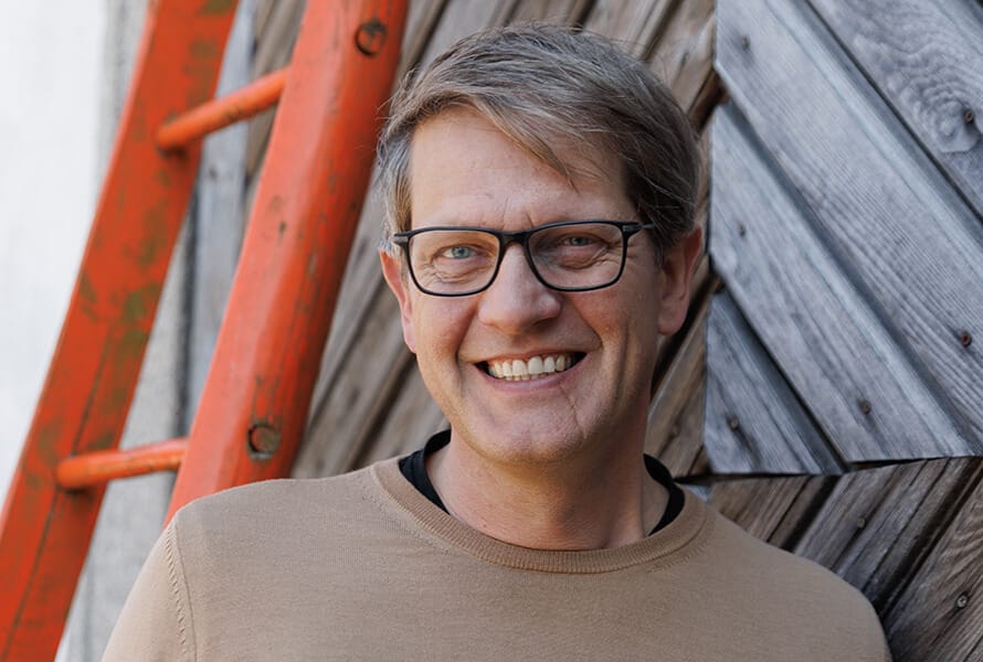 Norbert Eder vom Pankrazhofer lächelt in die Kamera. Er trägt eine Brille und ein beiges Oberteil. Im Hintergrund eine diagonale Holzfassade und eine rote Leiter.
