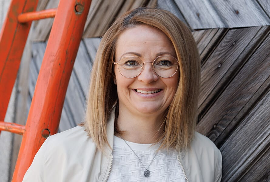 Susi Lumetzberger lächelt in die Kamera. Sie trägt eine helle Brille, eine weiße Jacke und eine Kette. Im Hintergrund eine diagonale Holzfassade und eine rote Leiter.